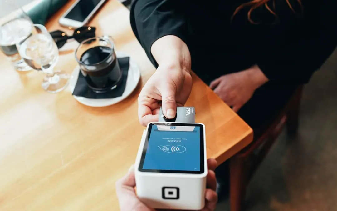 A hand holds a bank card to a payment terminal beside a coffee on a wooden table.