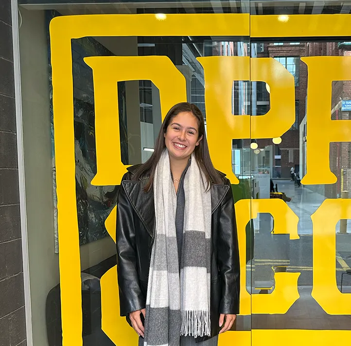 A smiling young woman in a black jacket and scarf stands in front of a large yellow DPR&Co sign.