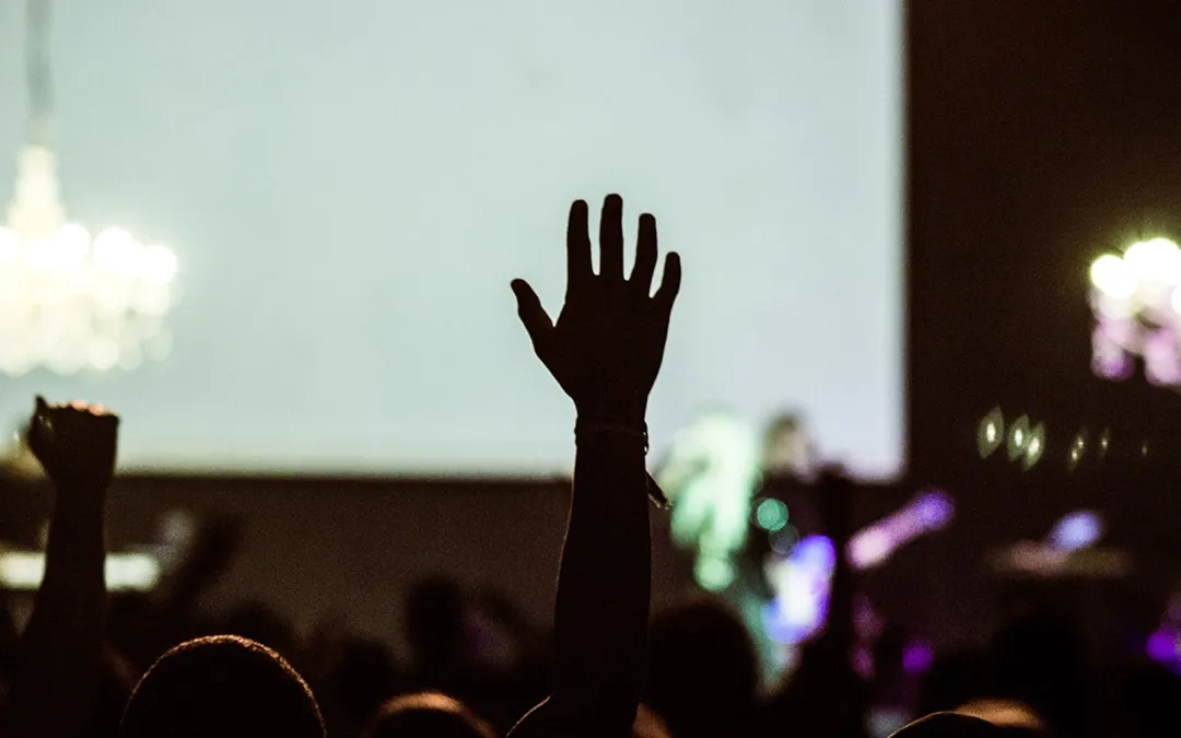 The silhouette of a raised hand in a dark crowd facing a bright screen.