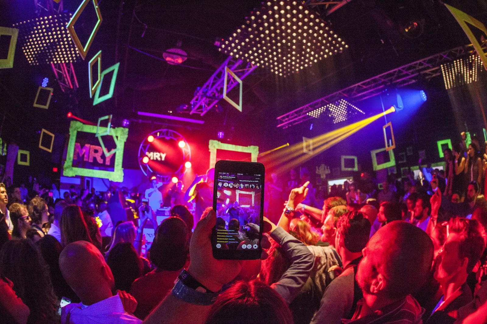 A crowd at a night event, with a person filming on their phone under neon green lights.