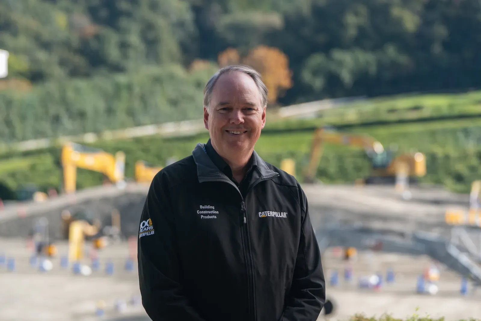 A smiling man in a Caterpillar jacket stands in front of a blurred construction site.