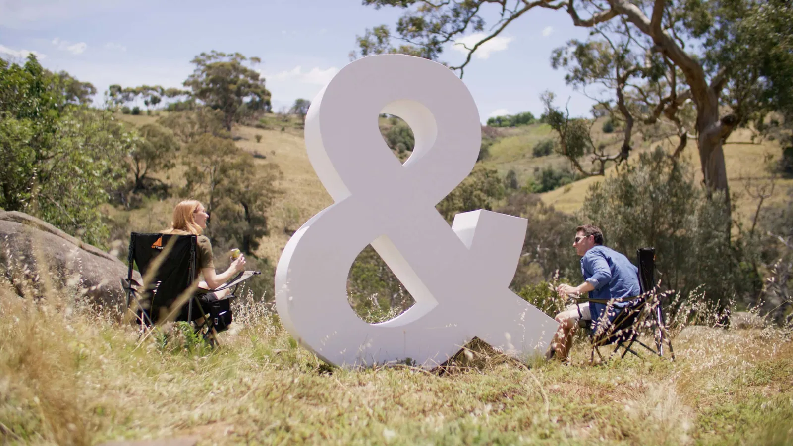 A large white ampersand stands on a grassy hillside while two people sit in folding chairs nearby.