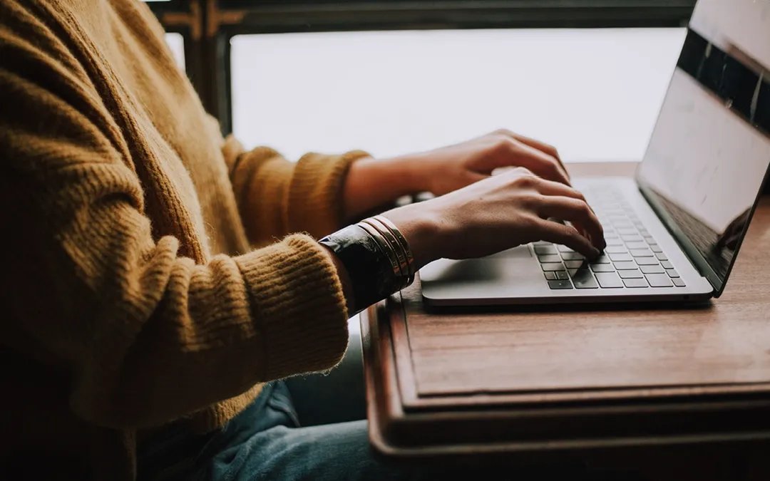 A person in a knitted jumper types on a laptop on a wooden table.