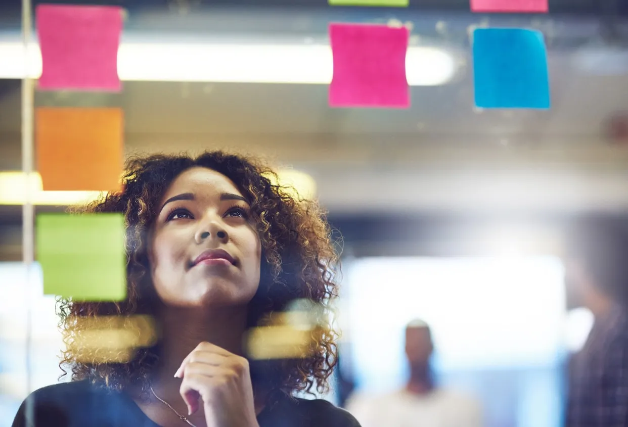 A woman looks thoughtfully upward in front of a glass wall covered in colourful sticky notes.
