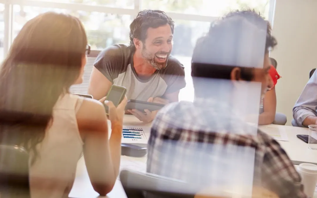 A group of colleagues laugh and talk around a table, seen through a glass partition.