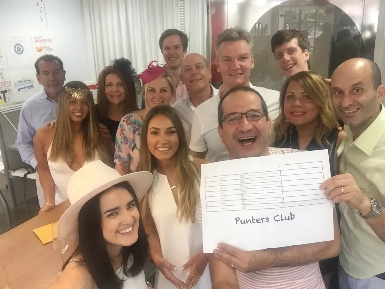 A group of smiling colleagues pose for a selfie, holding a handwritten sheet headed 'Punters Club'.