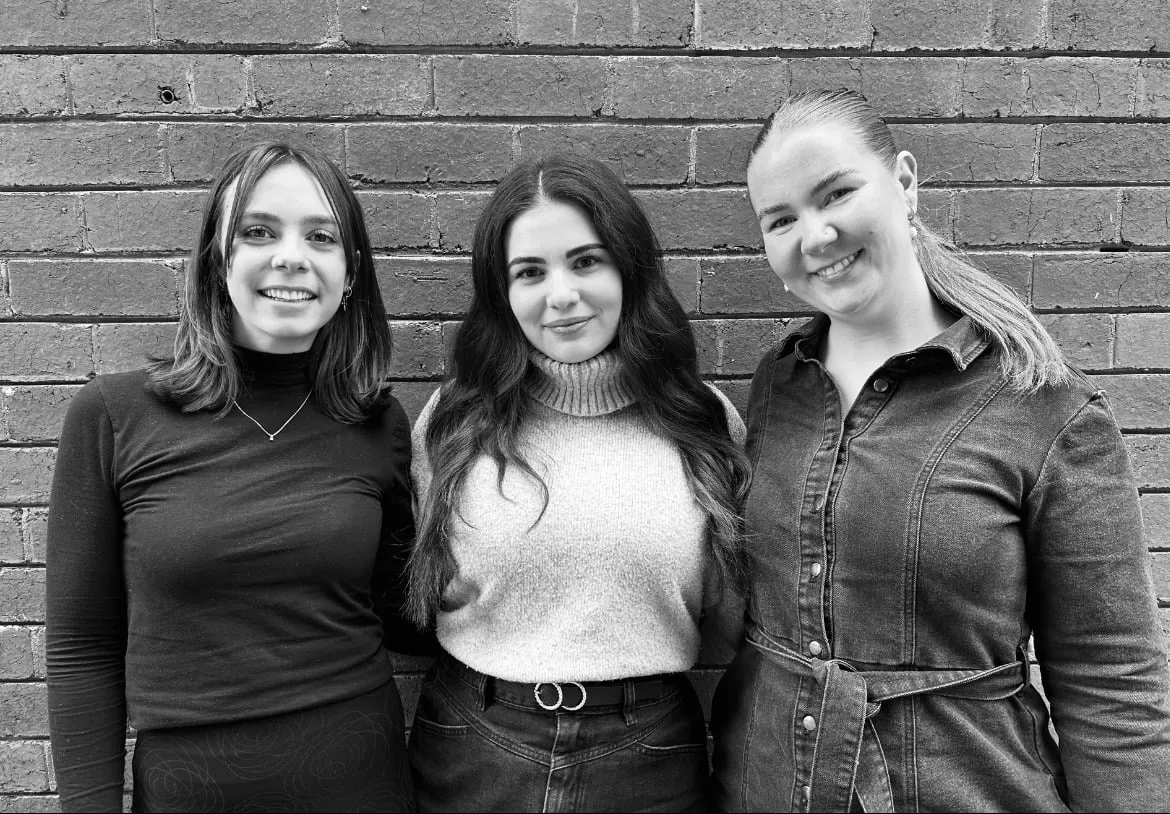 Three women stand smiling in front of a brick wall in a black-and-white photo.