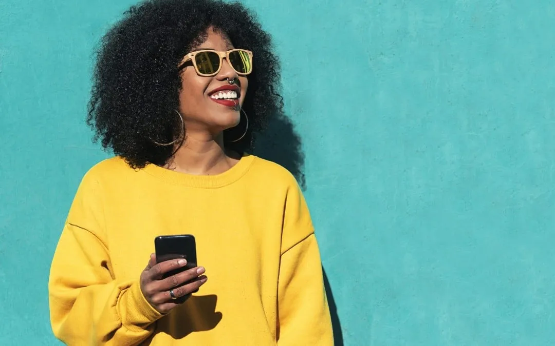 A smiling woman in yellow sunglasses and a yellow sweater holds a phone against a teal wall.