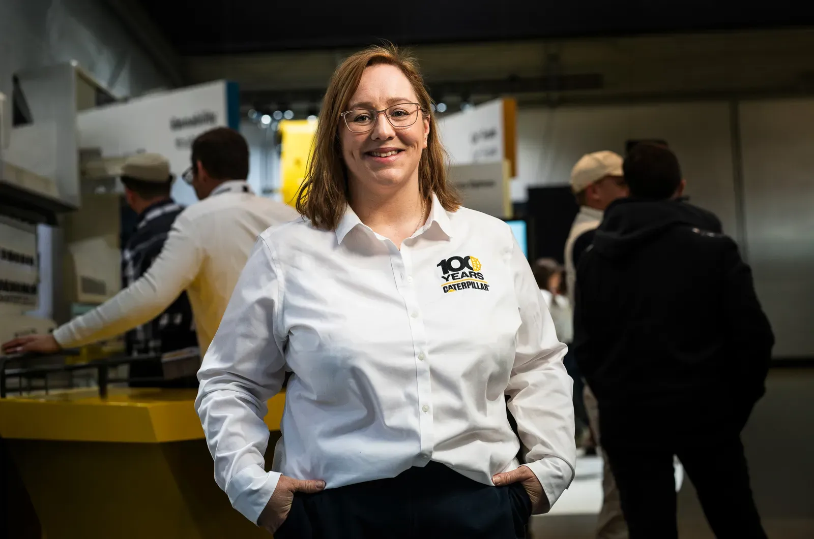 A smiling woman in a white Caterpillar 100 Years shirt stands in front of an exhibition floor.