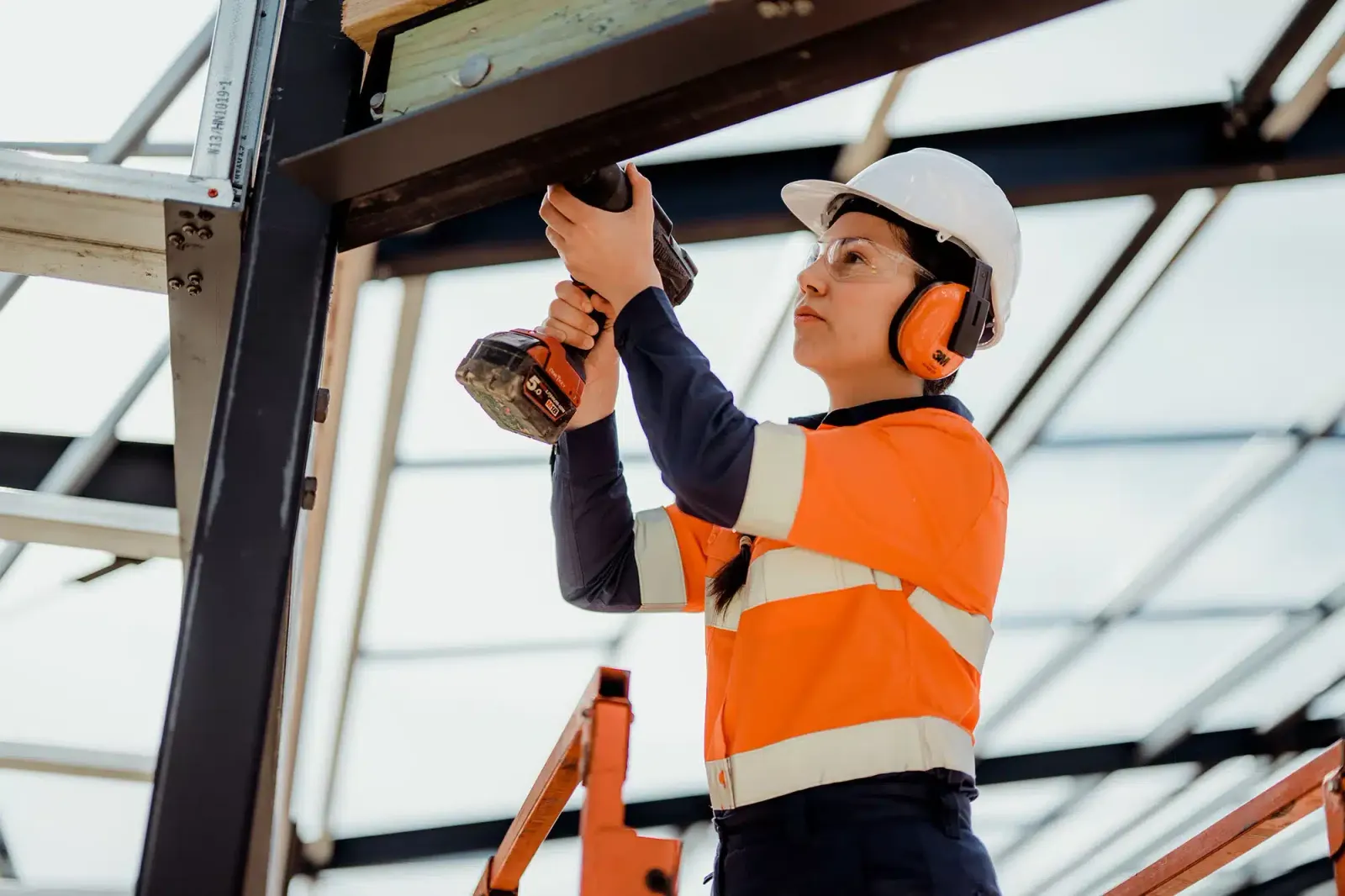 A worker in an orange hi-vis top, hard hat and ear protection uses a drill on a steel frame.