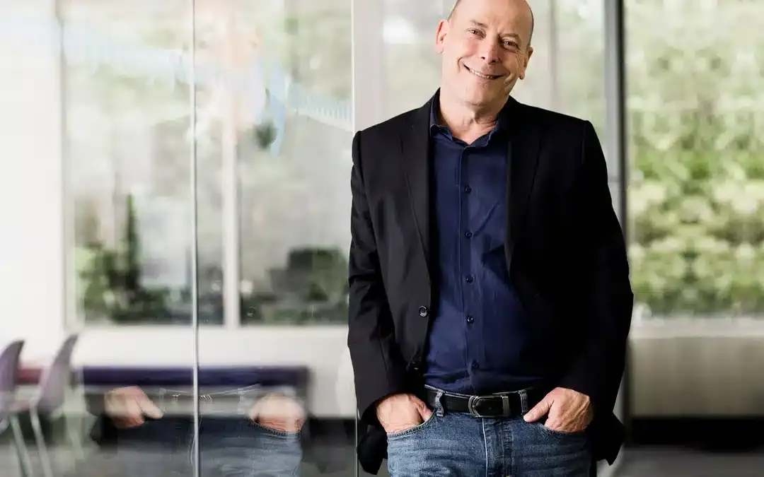 A smiling man in a dark jacket and jeans stands in a bright office with glass walls.