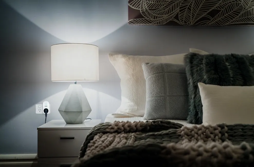 A lamp glows on a bedside table beside a neatly made bed.