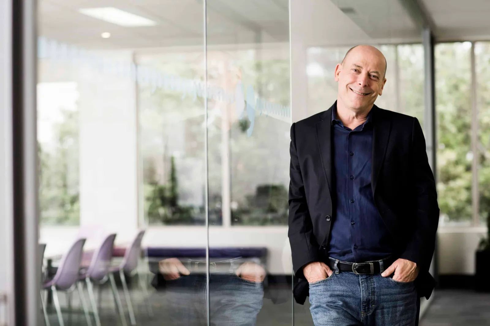 A smiling man in a dark jacket stands in a bright office with glass walls.