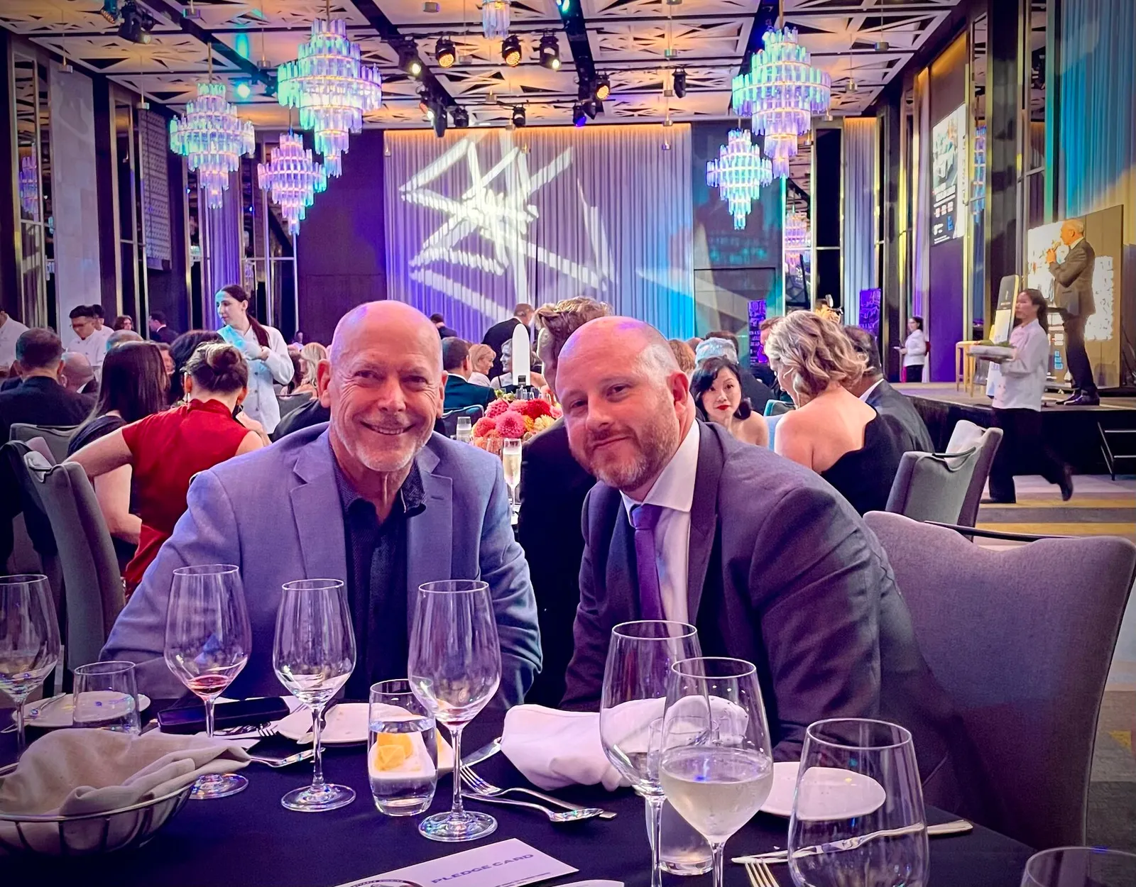 Two men in suits smile at a dinner table in a large event hall with chandeliers.