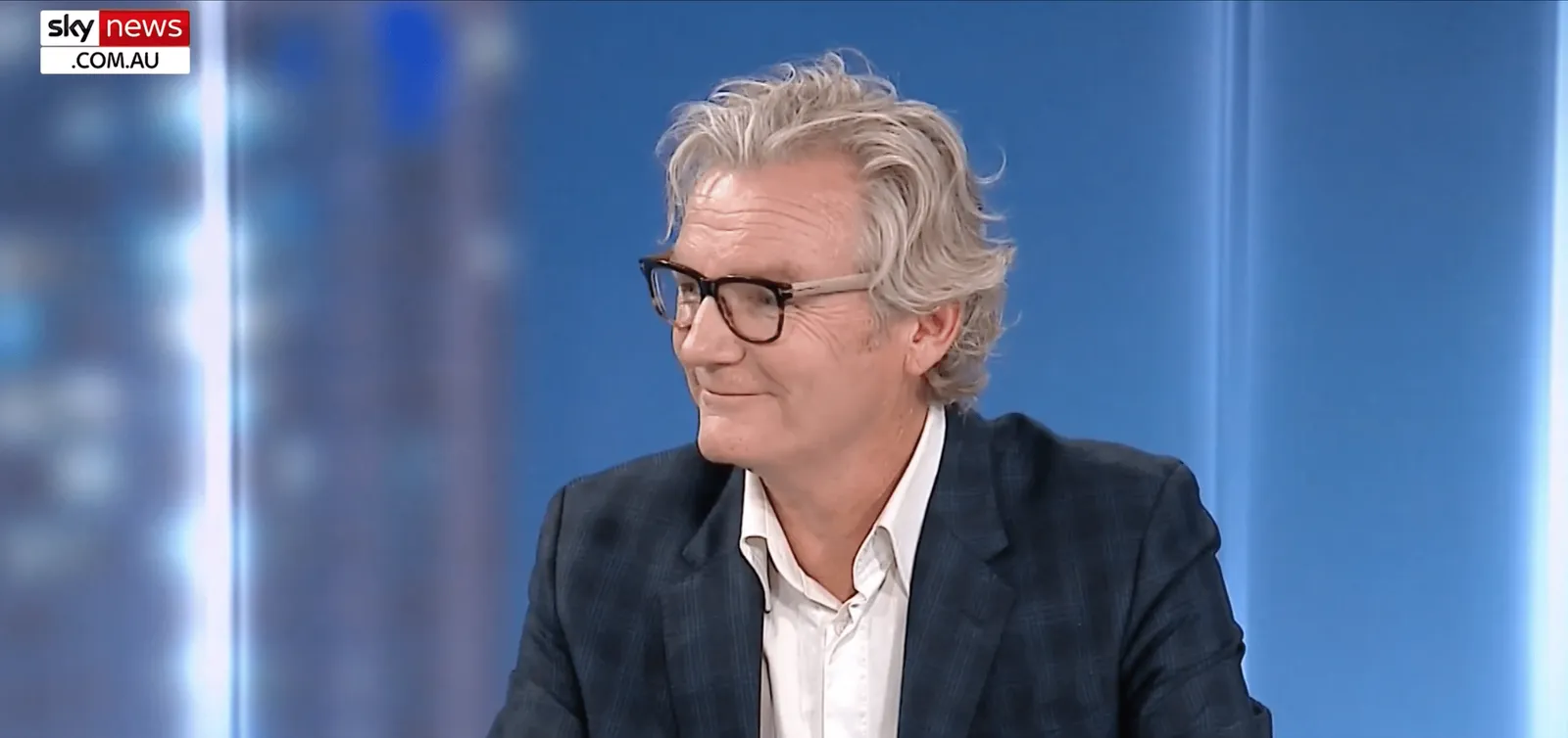 A man with grey hair and glasses on a television news set, looking to one side.