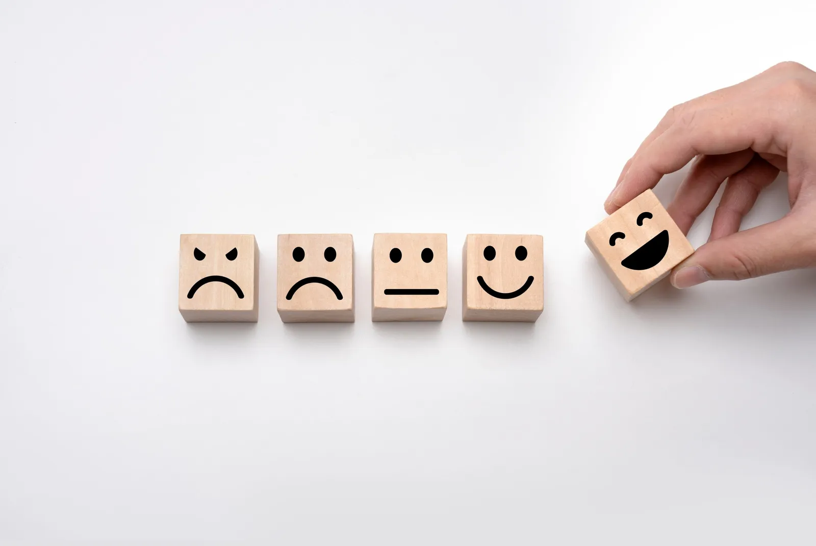 Five wooden blocks with faces from sad to happy, with a hand placing a smiling block on the end.