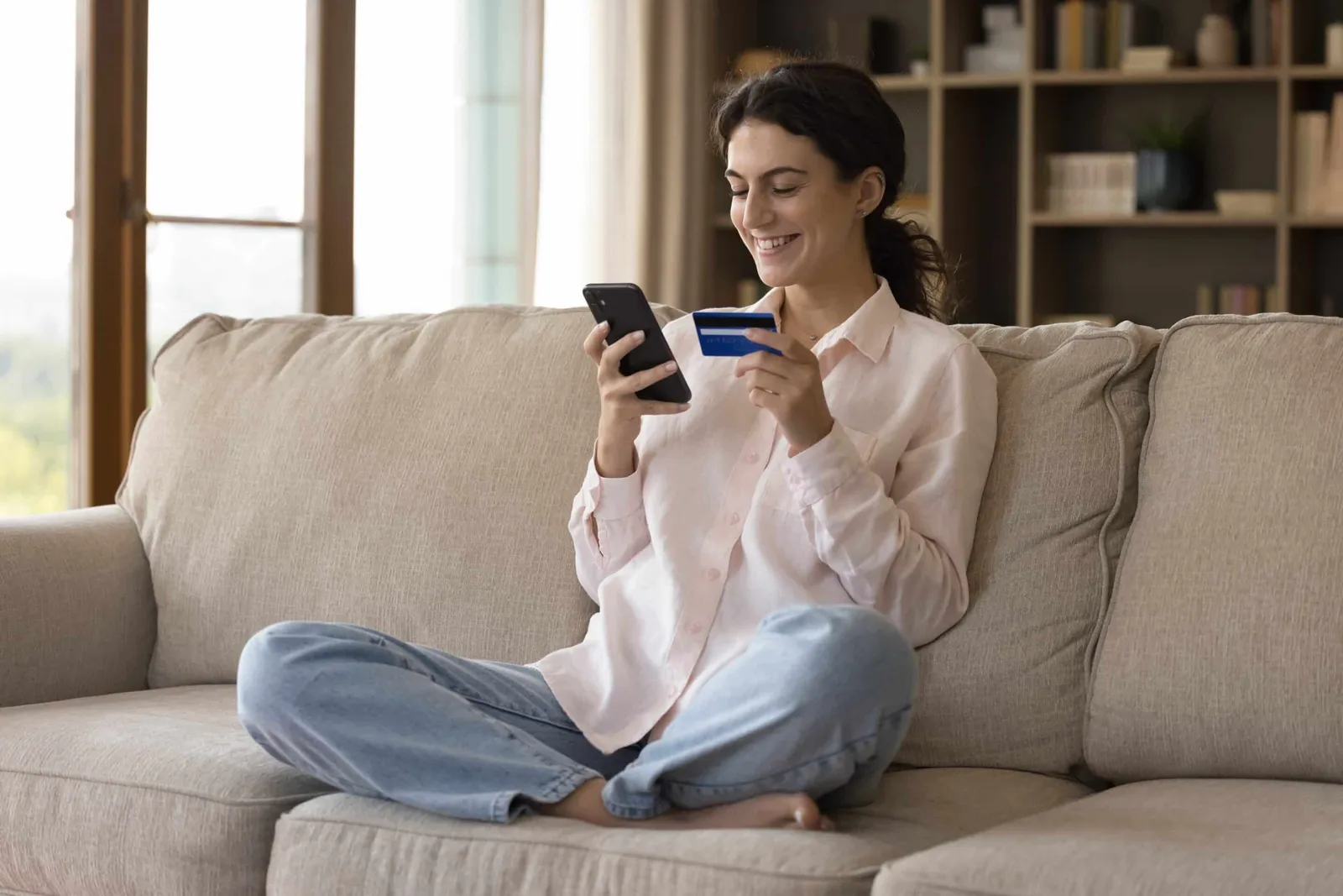 A woman sits cross-legged on a sofa, smiling at her phone while holding a bank card.