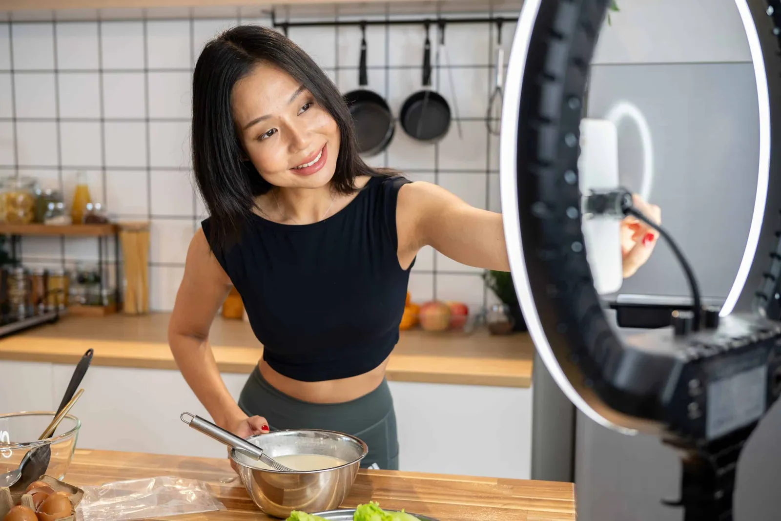 A woman films herself cooking in a kitchen, with a phone mounted on a ring light.