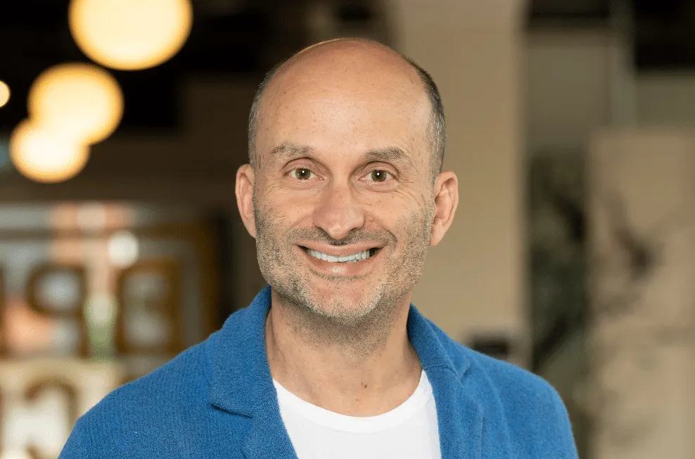 A smiling bald man in a blue jacket and white T-shirt in a café.