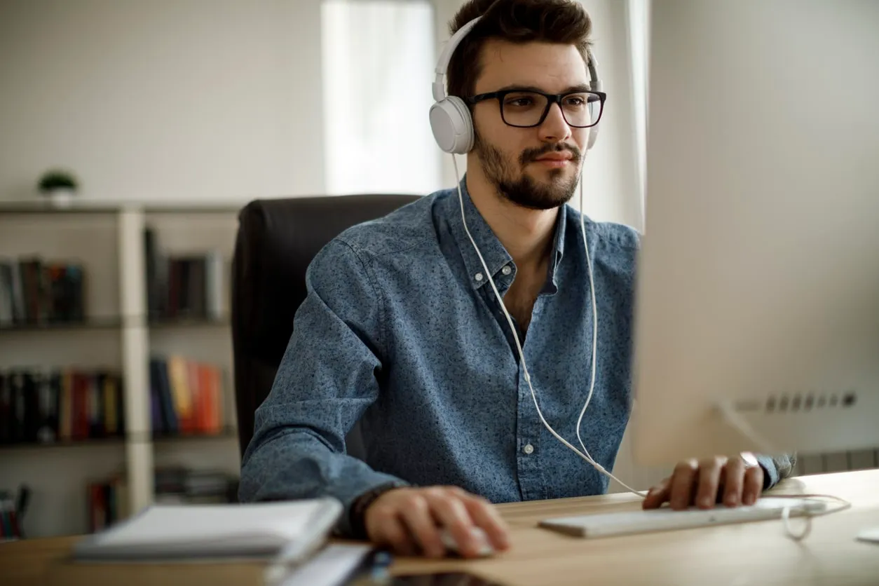 A man wearing headphones and glasses works at a desk in front of a computer.